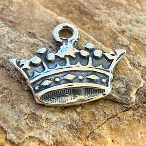 May include: A silver-toned metal charm shaped like a crown with a small circular loop at the top for attachment. The crown features a band with embossed diamond shapes and a series of rounded points along the upper edge with a metallic finish.