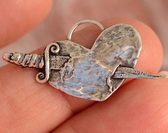 Puede incluir: Un colgante de plata en forma de corazón con una daga que lo atraviesa. El corazón es texturizado y la daga tiene un mango detallado.