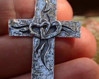 May include: A silver-colored cross pendant with a textured surface and a loop at the top. The cross features two intertwined hearts at the center and stylized flame-like designs. The pendant is held in a hand.