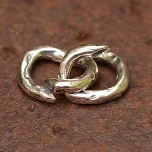 May include: Two interlocking silver rings, each with a textured, hammered finish. The rings are connected at a single point, creating a unique and modern design.
