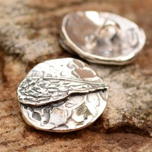 May include: Two silver earrings, each featuring a textured, round design with a raised wing motif. The wings are detailed with a textured surface.