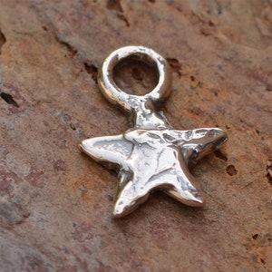 May include: A silver charm in the shape of a five-pointed star with a textured surface. The star has a loop at the top for attaching to a bracelet or necklace.