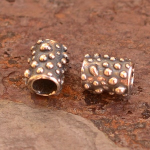 May include: Two cylindrical metal beads with a textured surface of small, rounded bumps. The beads have a dark, oxidized finish with highlights of a lighter, metallic color on the raised bumps. The beads are likely for jewelry making.