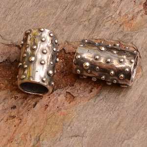 May include: Two silver metal beads with a textured surface. The beads are cylindrical and have a hammered finish with small raised dots.