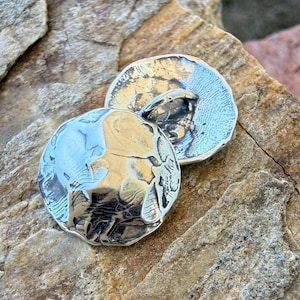 May include: Two silver-toned, hammered metal cufflinks with a textured surface. The cufflinks are round with a slightly irregular shape, reflecting light. They are displayed on a rough, tan-colored stone surface.