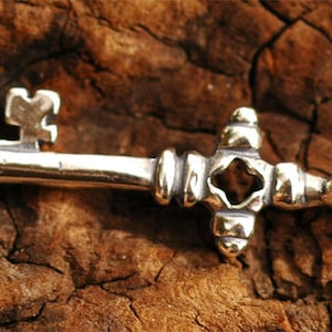 Peut inclure: Un charme de clé en argent avec un design orné. La clé a une grosse tête décorative et un long manche fin.