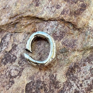 May include: A silver-toned ring with an open, irregular, and textured band. The metal surface features a polished finish with visible indentations and a gap between the two ends of the circular band, creating a distinct, non-uniform, and organic shape.