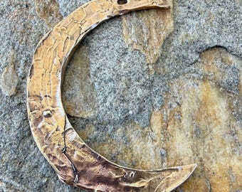 May include: A crescent moon pendant with a textured surface and a hole at the top for hanging. The pendant is a warm bronze color and rests on a gray and brown stone surface. The moon shape is a common symbol.