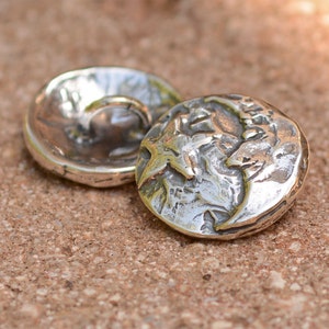 May include: Two silver-toned buttons with a textured surface. One button has a raised design of a face and the other button has a smooth surface with a loop on the top.