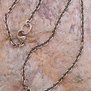 May include: A silver-colored chain necklace with an oval link design. The necklace has a heart-shaped clasp and is displayed on a textured, brown surface. The chain is made of metal and has a vintage or antique appearance.