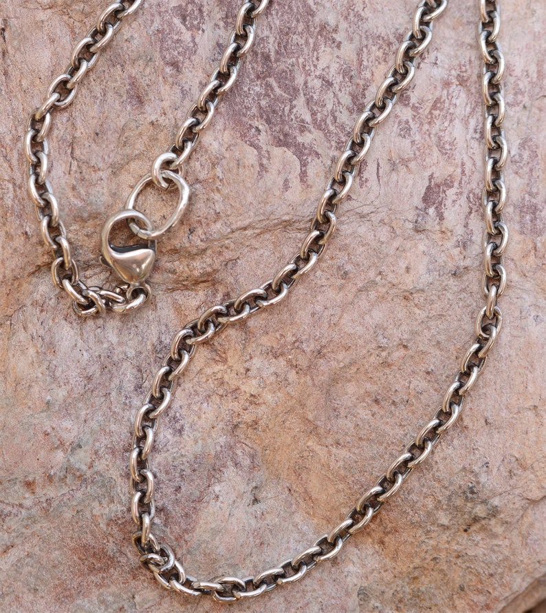May include: A silver-colored chain necklace with an oval link design. The necklace has a heart-shaped clasp and is displayed on a textured, brown surface. The chain is made of metal and has a vintage or antique appearance.