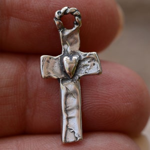 May include: A silver cross pendant with a heart in the center. The cross has a textured, hammered finish and a rope-like bail.
