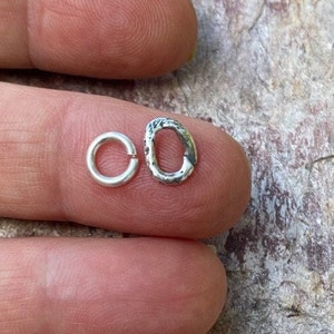May include: Two silver jump rings, one smooth and one textured, are shown on a finger.