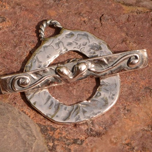 May include: Silver toggle clasp with a heart design and a textured circular piece. The clasp is designed for jewelry making.