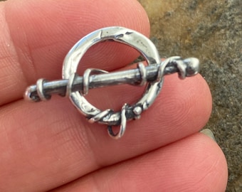May include: A silver toggle clasp featuring a circular ring with a hammered, uneven texture and a straight bar component. The bar is wrapped with a thin silver wire that coils around its length, creating a detailed, textured appearance.