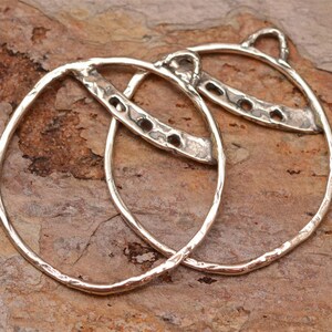 May include: Two silver hoop earrings with a hammered texture and a decorative cutout design. The hoops are slightly oval shaped and have a textured finish.