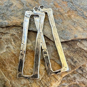 May include: A pair of silver-toned rectangular earrings with a textured, hammered appearance. The earrings are elongated, with a slightly irregular shape and a loop at the top for hanging. The background is a natural stone surface.