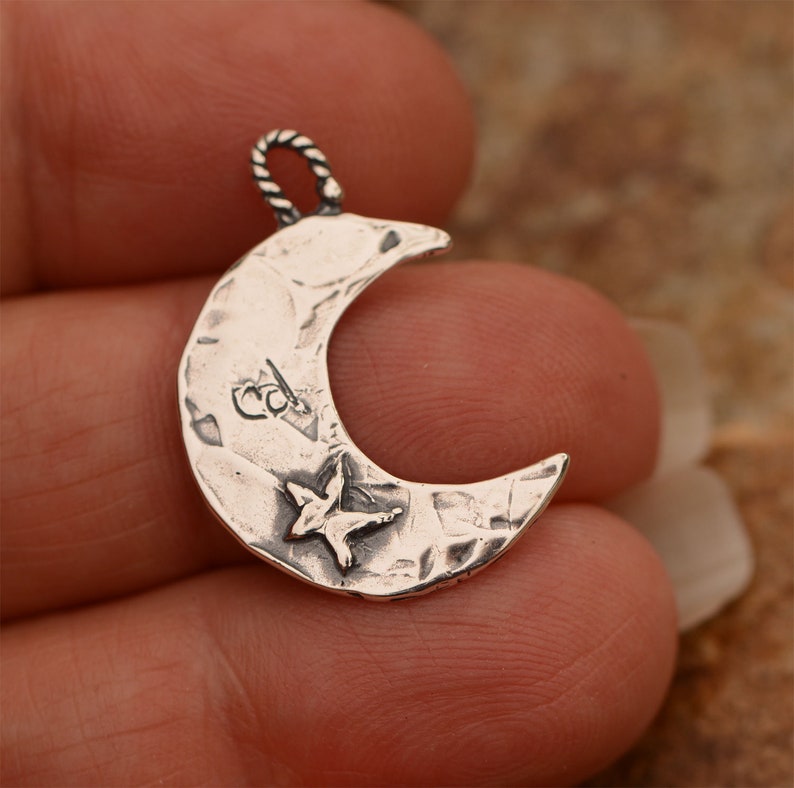 May include: A silver crescent moon charm with a textured surface and a small star in the center. The charm has a loop at the top for attaching to a necklace or bracelet.