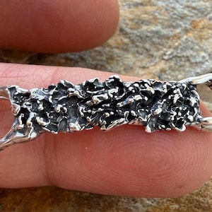 May include: A silver pendant with a textured, organic design. The rectangular piece has a rough, uneven surface and rounded loops on each end. The pendant is held between fingers, showcasing its intricate details and metallic sheen.