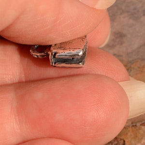 May include: Close-up of a silver charm in the shape of a closed book. The charm has a detailed, textured surface and a small loop for attaching to a bracelet or necklace.