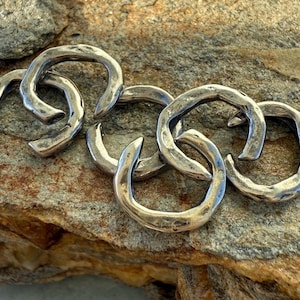 May include: Close-up of several silver-colored, horseshoe-shaped metal links. The links are interlocked and have a textured, hammered appearance. They rest on a rough, tan and gray stone surface.
