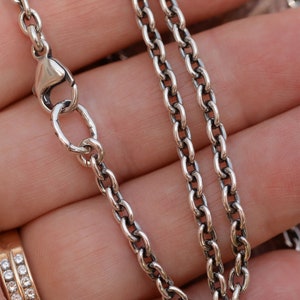 May include: Close-up of a silver-colored chain necklace with an oval link design. The necklace is shown against a neutral background, with a lobster clasp closure. The chain is a classic jewelry piece.