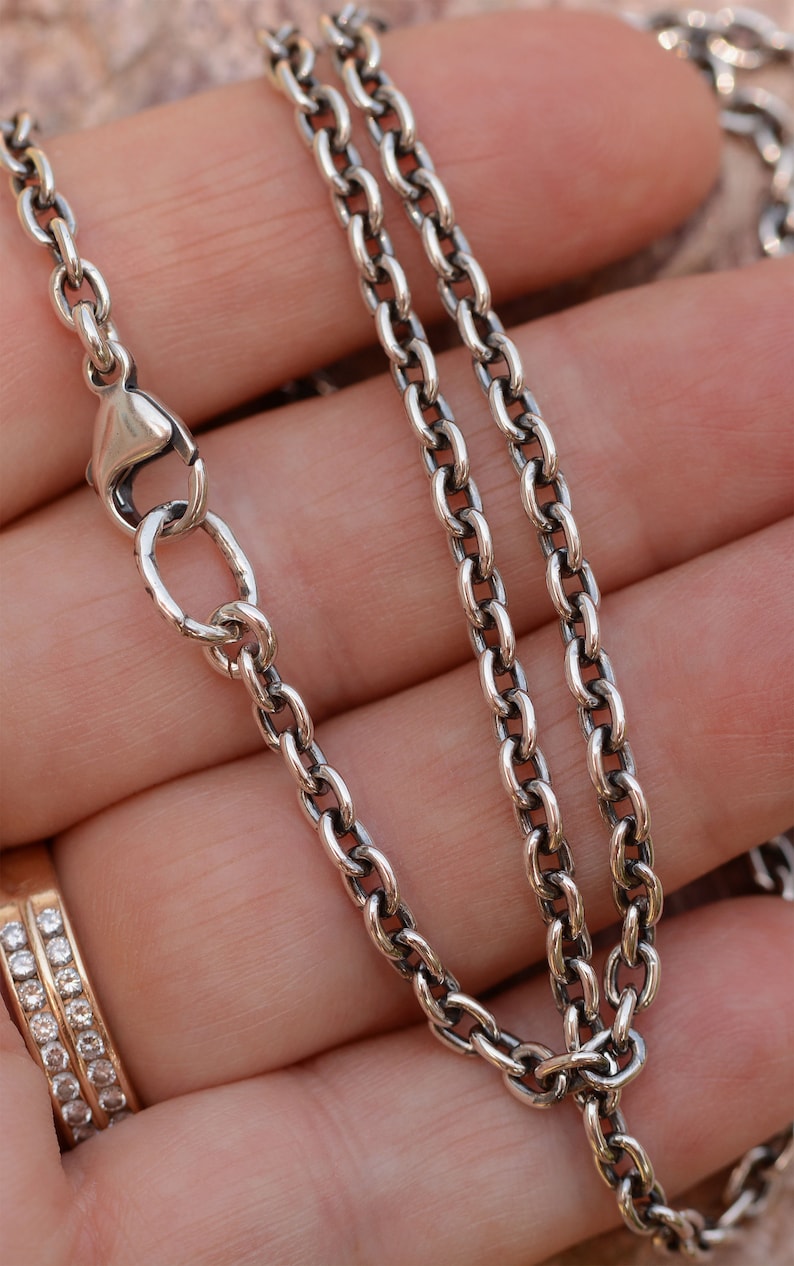 May include: Close-up of a silver-colored chain necklace with an oval link design. The necklace is shown against a neutral background, with a lobster clasp closure. The chain is a classic jewelry piece.