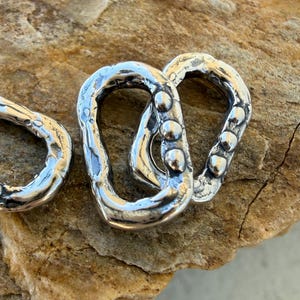May include: Two interlocking silver-toned metal links with a hammered, irregular texture. The links feature a series of small, raised circular bumps along one side, creating a rugged, organic appearance with a polished, reflective finish.