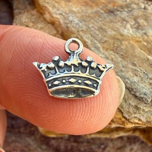 Puede incluir: Un peque&ntilde;o dije de metal en tono plateado con forma de corona y un lazo circular en la parte superior para sujetarlo. La corona presenta una banda detallada con patrones de diamantes en relieve y puntas redondeadas en el borde superior.