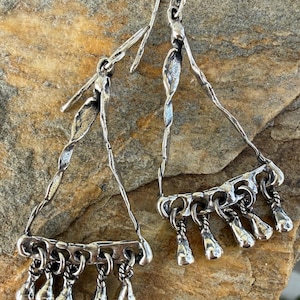May include: A pair of silver-toned earrings. Each earring has a triangular frame with a twisted design and dangling teardrop-shaped charms. The earrings are displayed on a textured, tan-colored surface.