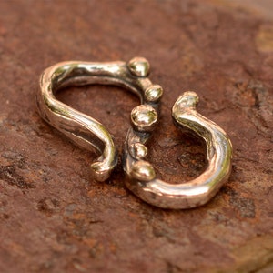 K&ouml;nnte beinhalten: Silberfarbener Metallverschluss mit einem strukturierten, organischen Design. Der Verschluss hat eine abgerundete, offene Schlaufe auf der einen Seite und eine geschlossene Schlaufe auf der anderen. Die geschlossene Schlaufe hat eine Reihe kleiner, runder Perlen entlang der Kante.
