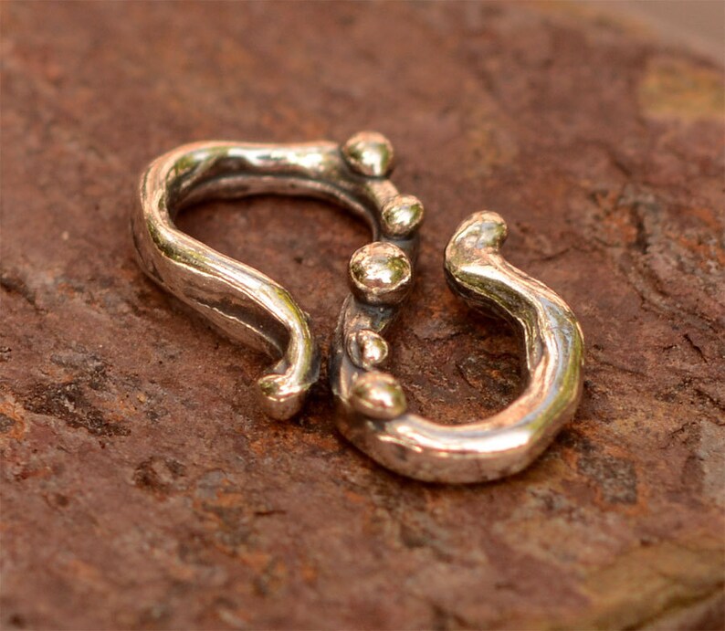 K&ouml;nnte beinhalten: Silberfarbener Metallverschluss mit einem strukturierten, organischen Design. Der Verschluss hat eine abgerundete, offene Schlaufe auf der einen Seite und eine geschlossene Schlaufe auf der anderen. Die geschlossene Schlaufe hat eine Reihe kleiner, runder Perlen entlang der Kante.
