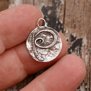 May include: A silver charm with a textured surface and a swirl design. The charm has a loop at the top for attaching to a necklace or bracelet.