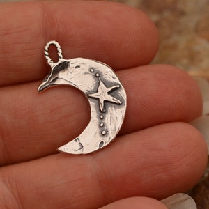 May include: A silver crescent moon charm with a five-pointed star in the center. The moon has a textured surface and a small loop at the top for attaching to a necklace or bracelet.