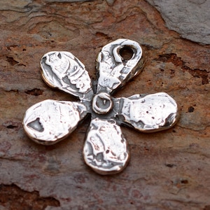 May include: A silver charm in the shape of a flower with five petals. The charm has a textured surface and a hole at the top for attaching to a chain or bracelet.