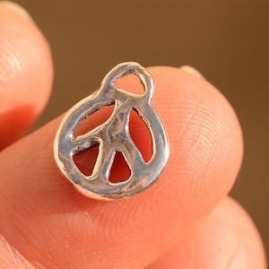 May include: A small, silver-colored peace symbol charm. The charm is held between two fingers and has a rounded shape with the iconic peace sign design. The metal has a slightly textured appearance.