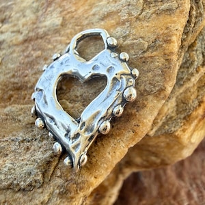 May include: A silver-toned heart-shaped pendant with a textured, hammered surface and a series of small, raised spherical beads along the outer edge. The center of the heart is open, and a small loop is positioned at the top for attachment.