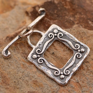 May include: Silver toggle clasp with a square frame design featuring intricate scrollwork and a hammered texture.
