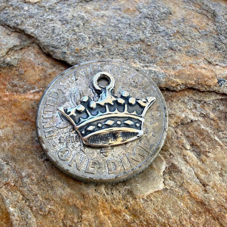 Puede incluir: Un dije de corona de metal plateado con un peque&ntilde;o lazo en la parte superior para sujetarlo, que descansa directamente sobre una moneda de diez centavos de los Estados Unidos. La corona presenta una banda detallada con diamantes en relieve.