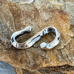 May include: A silver-toned metal S-hook connector featuring a hammered, textured surface with a polished finish. The hook has a curved, open-ended design with rounded tips, showcasing a reflective metallic sheen and a rugged, hand-forged appearance.