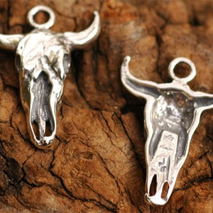 May include: Two silver bull skull charms with a detailed design. The charms have a loop at the top for attaching to a necklace or bracelet.