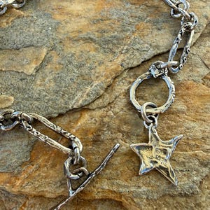 May include: A silver-toned chain bracelet featuring a toggle clasp and a five-pointed star charm. The chain consists of elongated, textured oval links with a distressed, metallic finish. The star charm hangs from a circular link near the toggle.