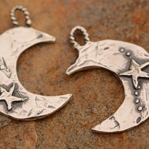May include: Two silver crescent moon charms with a star design. One charm has a star with a textured surface and the other has a star with a smooth surface.