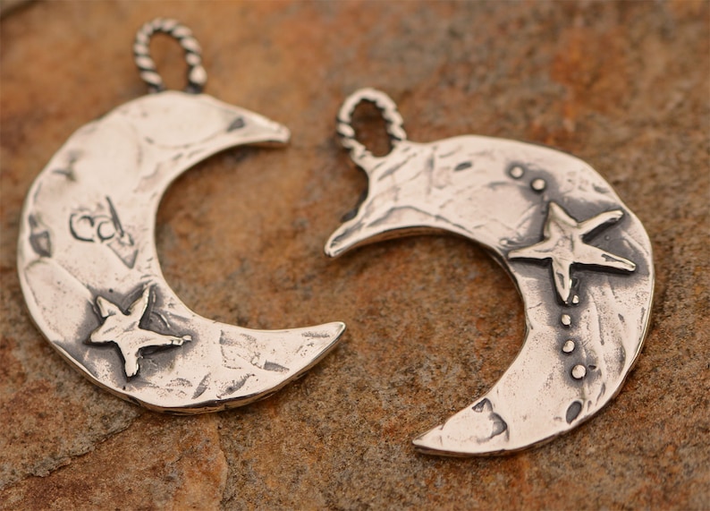 May include: Two silver crescent moon charms with a star design. One charm has a star with a textured surface and the other has a star with a smooth surface.
