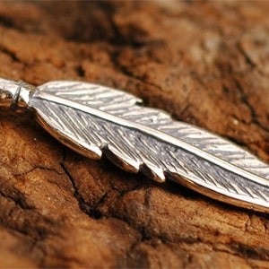 May include: A silver-colored feather pendant with intricate detailing. The pendant has a loop at the top for hanging and is set against a textured, brown wooden surface. The feather design is detailed and realistic.