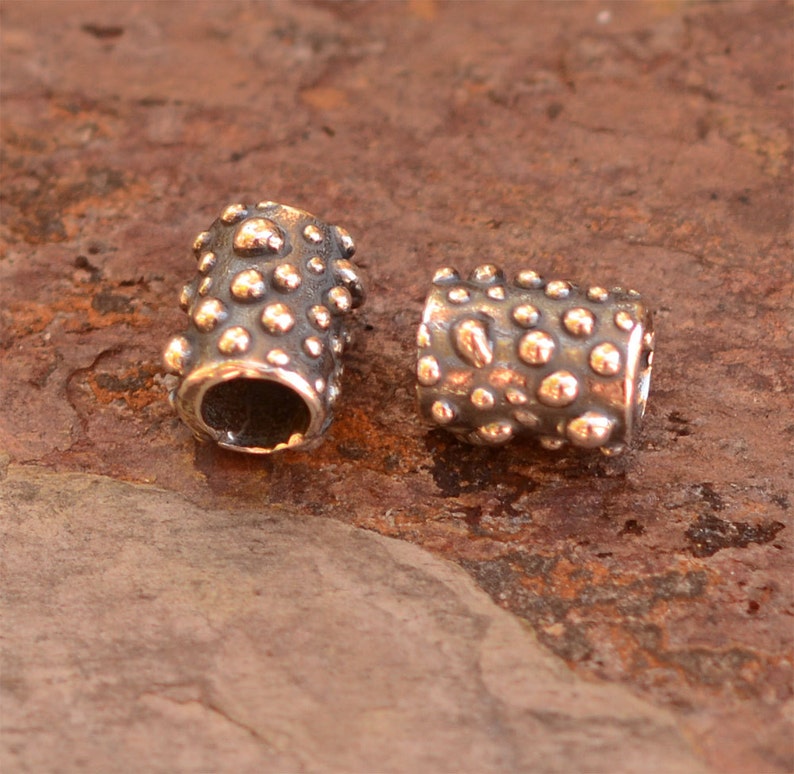 May include: Two cylindrical metal beads with a textured surface of small, rounded bumps. The beads have a dark, oxidized finish with highlights of a lighter, metallic color on the raised bumps. The beads are likely for jewelry making.