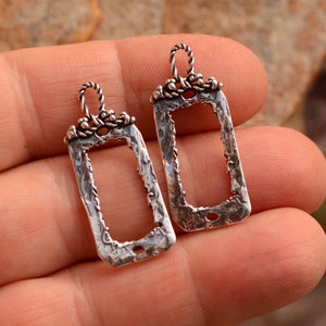 May include: Two silver-toned metal rectangular frame pendants with a textured finish and a twisted wire loop at the top.