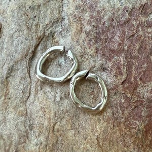 May include: Two silver-colored, open-hoop earrings with an organic, textured design. The earrings are circular with a small gap at the top, and they rest on a rough, stone-like surface. The earrings have a hammered appearance.