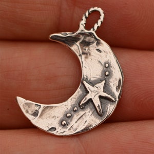 May include: A silver crescent moon charm with a five-pointed star and small dots. The charm has a textured surface and a loop at the top for attaching to a necklace or bracelet.