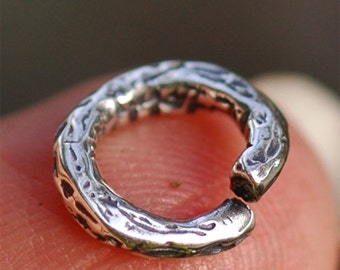 Puede incluir: Un anillo de salto de plata con una superficie texturizada. El anillo está abierto y listo para ser utilizado para la fabricación de joyas.